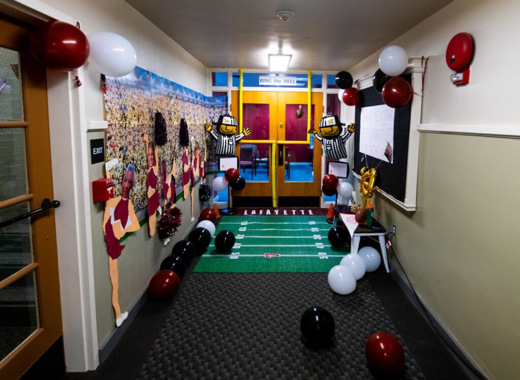 As part of the 2024 office decorating contest a hallway is shown with a green endzone on the floor, balloons in maroon and white, yellow goal posts at the door and leopard print banners on the wall.