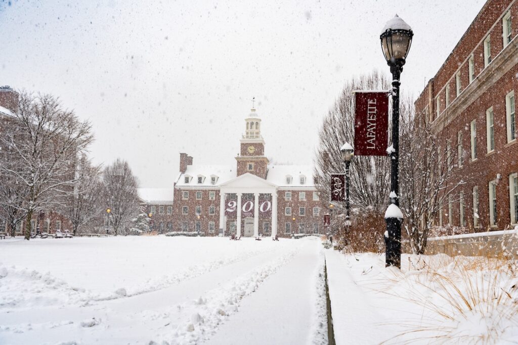 A snowy scene shows snow falling and blanketing the ground of Anderson Courtyard. The scene is framed with lightposts with Lafayette and 200 banners one them. A giant "200" Bicentennial banner hangs on Watson Hall.