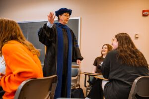 Prof. John Shaw in his cap and gown speaks with a room of students.