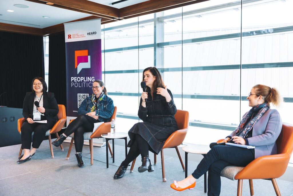 Four panelists sit on a stage while one speaks into a microphone. Caroline Lee is the second from left.